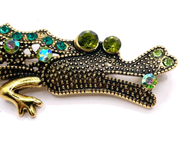 May include: A gold-toned metal alligator brooch with green and iridescent rhinestones. The alligator is in a walking position with its head turned to the side. The brooch is detailed with a textured pattern and has a shiny finish.