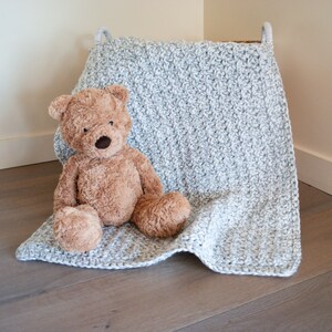 May include: A gray crocheted baby blanket with a teddy bear sitting on top of it.