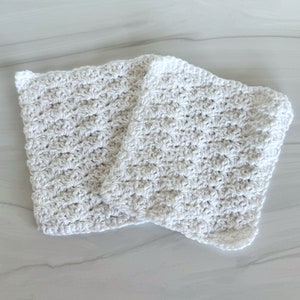 May include: Two white crocheted dishcloths, made with a textured stitch pattern, are folded together on a white marble surface.