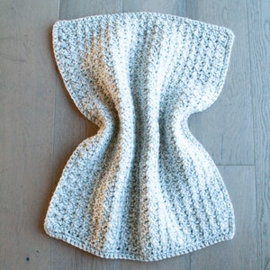 May include: A gray and white crocheted blanket with a textured pattern. The blanket is folded in half and laid flat on a wooden surface.