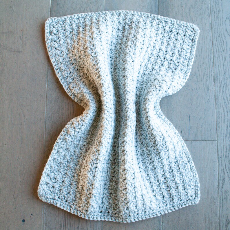 May include: A gray and white crocheted blanket with a textured pattern. The blanket is folded in half and laid flat on a wooden surface.
