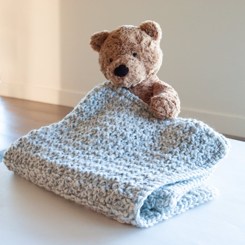 May include: A light blue crocheted baby blanket with a brown teddy bear peeking out from under it.