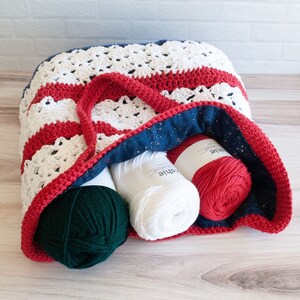 May include: A handmade crochet tote bag in red, white, and blue, filled with yarn. The bag features a striped pattern and a red handle. Inside, there are several balls of yarn in white, red, and green. The bag's interior is lined with blue fabric.