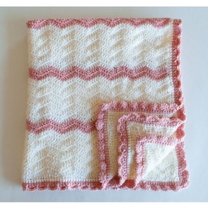 May include: A white crochet baby blanket with a pink crocheted border. The blanket has a wavy pattern and is folded in half. The text "CROCHET BABY BLANKET PATTERN" is at the top of the image and "WWW.CRAFTINGEACHDAY.COM" is at the bottom.
