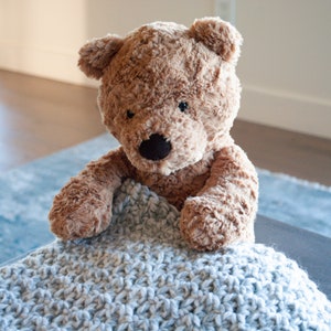 May include: A brown teddy bear with black eyes and a black nose is nestled in a light blue crocheted blanket.