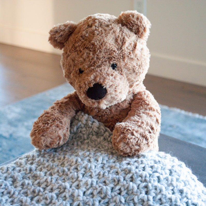 May include: A brown teddy bear with black eyes and a black nose is nestled in a light blue crocheted blanket.