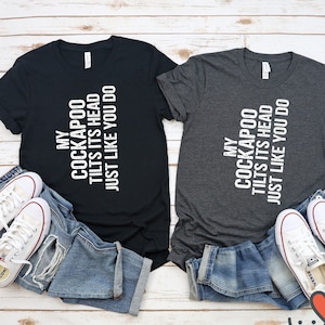May include: Two t-shirts with the text "My Cockapoo Tilts Its Head Just Like You Do" printed in white on a black and a gray shirt. The shirts are laid out on a white wooden surface with a pair of white sneakers and blue jeans on each side.