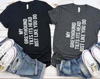May include: Two t-shirts with the text "My Greyhound Tilts Its Head Just Like You Do" in white on a black and a grey shirt. The shirts are laid out on a white wood surface with a pair of white sneakers and blue jeans on each side.