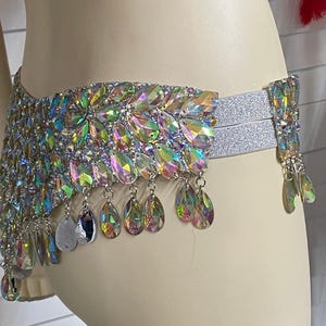 Pode incluir: Um cinto de cintura com cristais iridescentes em forma de l&aacute;grima dispostos num padr&atilde;o floral denso. O cinto inclui faixas el&aacute;sticas prateadas e pendentes de cristal que balan&ccedil;am na borda inferior, criando um efeito cintilante.