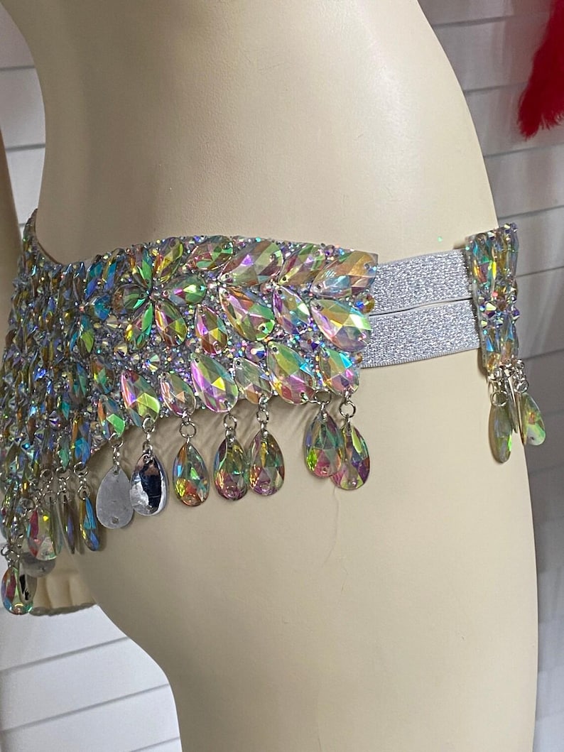 Pode incluir: Um cinto de cintura com cristais iridescentes em forma de l&aacute;grima dispostos num padr&atilde;o floral denso. O cinto inclui faixas el&aacute;sticas prateadas e pendentes de cristal que balan&ccedil;am na borda inferior, criando um efeito cintilante.