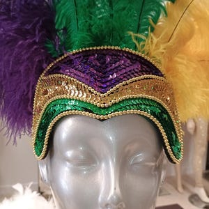 Pode incluir: Um adereço de cabeça de Mardi Gras com penas e lantejoulas roxas, verdes e douradas. O adereço é decorado com contas douradas e tem uma forma curva.