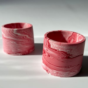 Puede incluir: Dos pequeñas macetas cilíndricas con un diseño veteado. Las macetas son principalmente rosas con remolinos blancos, creando un patrón único. El interior de las macetas es de color rosa liso. Las macetas están sobre un fondo blanco.