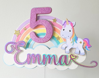 May include: A pink glitter number 5 cake topper with a rainbow and a unicorn. The topper also includes the name "Emma" with a pink heart.
