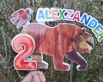 May include: A cake topper featuring a brown bear, a red cardinal, and the number 2. The bear is walking and has a brown and white striped pattern. The cardinal is flying and has red and white feathers. The number 2 is red and has a gold outline. The name "ALEXZANDER" is written in rainbow letters above the bear.