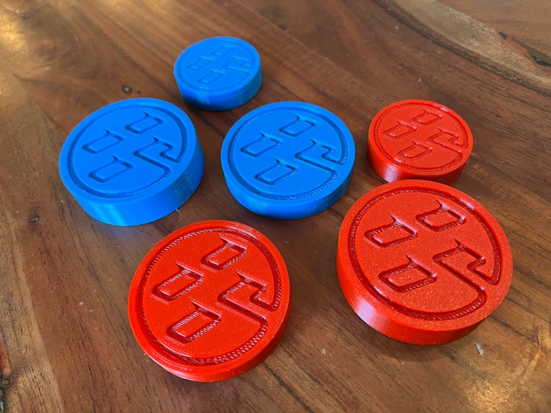 May include: Six round, 3D printed game pieces. Three are blue with a white symbol and three are orange with a white symbol.