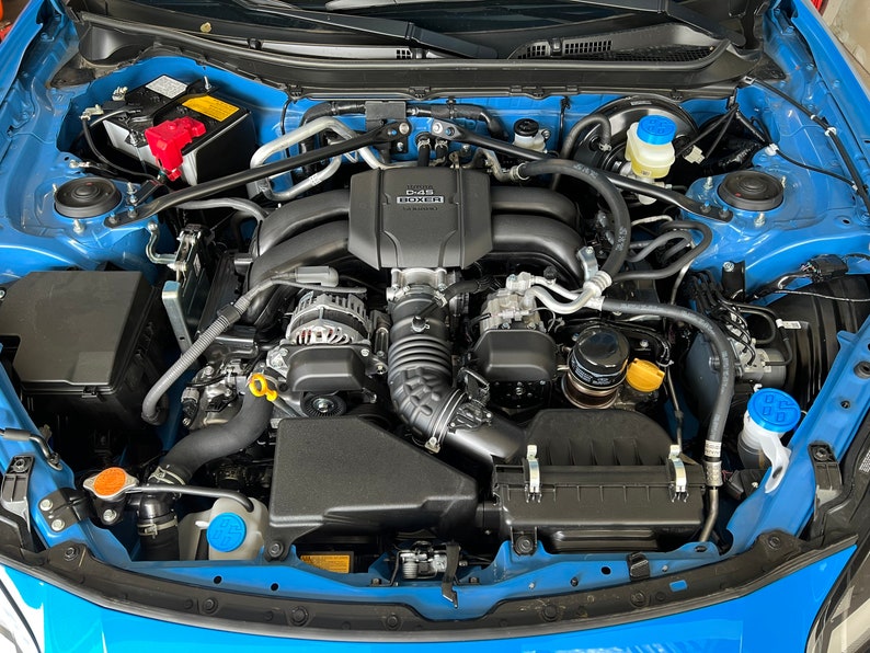 May include: A close-up view of a blue car engine with black hoses and a black air filter. The engine is clean and well-maintained. The engine has a black plastic cover with the text "D4-S" printed on it.