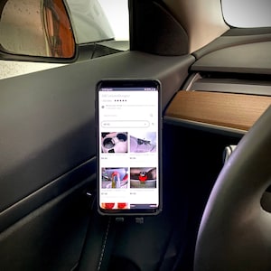 May include: A black smartphone with a white screen displaying a product listing from the website "ABC Custom Designs" with a five-star rating. The listing shows various products, including sunglasses, a bottle opener, and a bottle stopper. The phone is mounted in a car.