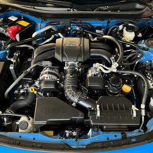 May include: Close-up view of a blue car engine with a black air filter, a black engine cover, and a black alternator. The engine cover has the text "2.0L BOXER" printed on it.
