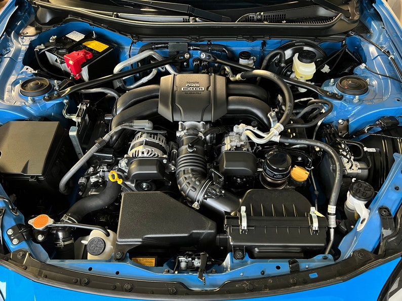 May include: Close-up view of a blue car engine with a black air filter, a black engine cover, and a black alternator. The engine cover has the text "2.0L BOXER" printed on it.