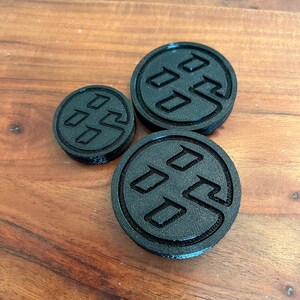 May include: Three black 3D printed round coasters with a raised design of a stylized logo. The logo is a circle with three overlapping squares inside.