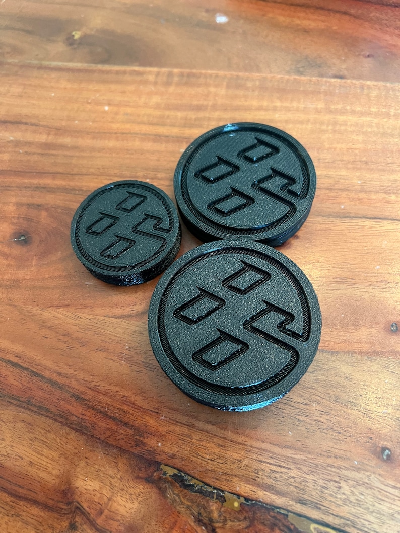 May include: Three black 3D printed round coasters with a raised design of a stylized logo. The logo is a circle with three overlapping squares inside.
