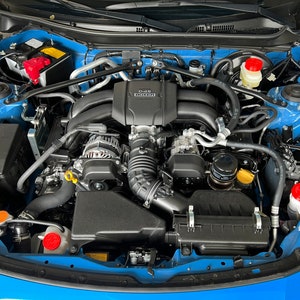 May include: A close-up view of a blue car engine with a black engine cover. The engine cover has the text "D-4S" and "SUBARU" printed on it. The engine is clean and well-maintained.
