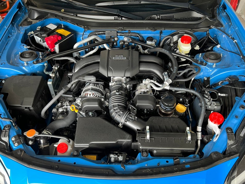 May include: A close-up view of a blue car engine with a black engine cover. The engine cover has the text "D-4S" and "SUBARU" printed on it. The engine is clean and well-maintained.