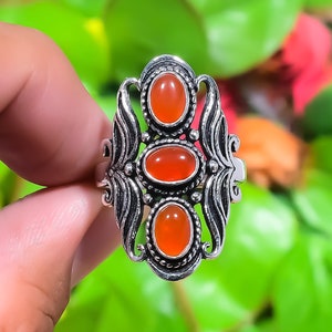 Puede incluir: Un anillo de plata con tres piedras preciosas ovaladas de color rojo anaranjado. El anillo tiene un diseño de filigrana ornamentado con elementos decorativos en los laterales. Las piedras están engastadas en biseles individuales, sobre un fondo verde borroso.