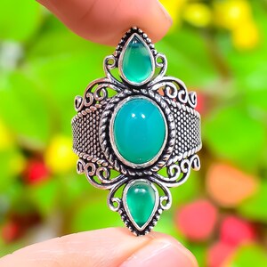 Peut inclure: Une bague en argent ornée d'une grande pierre verte ovale, flanquée de deux petites pierres vertes en forme de goutte. La bague présente un design filigrané orné et une bande texturée. La bague est tenue par les doigts d'une personne.