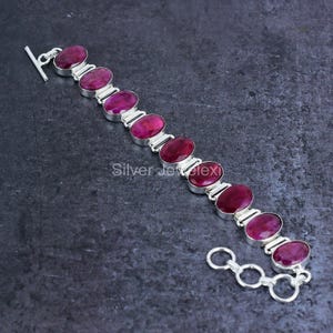 May include: A silver bracelet featuring oval-shaped, deep red gemstones. The gemstones are set in individual silver settings and connected by silver links. The bracelet has a toggle clasp closure. The jewelry is displayed on a dark gray surface.
