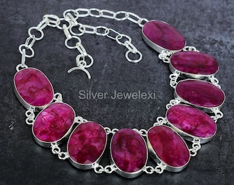 May include: A silver necklace featuring oval-shaped, deep pink gemstones. The gemstones are set in a silver bezel and connected by a silver chain. The necklace has a hook clasp. The background is a dark gray.