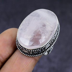May include: A silver ring with a large, oval rose quartz gemstone. The gemstone is a pale pink color and has a smooth, polished surface. The ring has a detailed, textured silver band. The text "Silver Jewelexi" is visible on the ring.