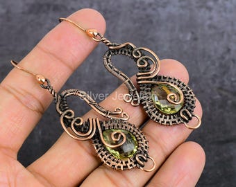 May include: A pair of dangle earrings featuring teardrop-shaped light green gemstones encased in intricate, dark-toned copper wire wrapping. The metalwork displays a complex pattern of coils and loops, finished with simple copper ear hooks.