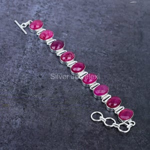 Op de afbeelding: Een zilveren armband met een ketting en een sluiting. De armband is versierd met ovale roze edelstenen.