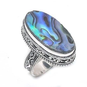 Puede incluir: Anillo ovalado de plata con una piedra de concha de abulón iridiscente en tonos azules y verdes. La banda presenta intrincados diseños de volutas y un borde decorativo.