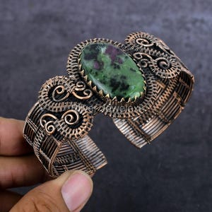 Pode incluir: Uma pulseira tipo bracelete feita de fio de cobre oxidado com padrões entrelaçados e espirais complexas. A peça central é uma grande pedra de zoisite oval com cores verdes e pretas proeminentes e pequenas inclusões em vermelho magenta.
