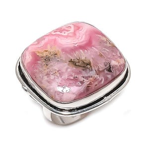 May include: A square-shaped ring featuring a large, polished gemstone with a swirling pattern of pink, white, and brown hues. The gemstone is set in a silver-colored metal band. The ring has a modern, elegant design.