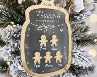 Puede incluir: Un adorno navideño en forma de tarro con borde dorado y centro gris oscuro. Incluye el texto 2026, Nanna's PERFECT BATCH, y cinco iconos dorados de hombrecitos de jengibre etiquetados como Jesse, Chloe, Ryan, Hayley y Arlo con copos.