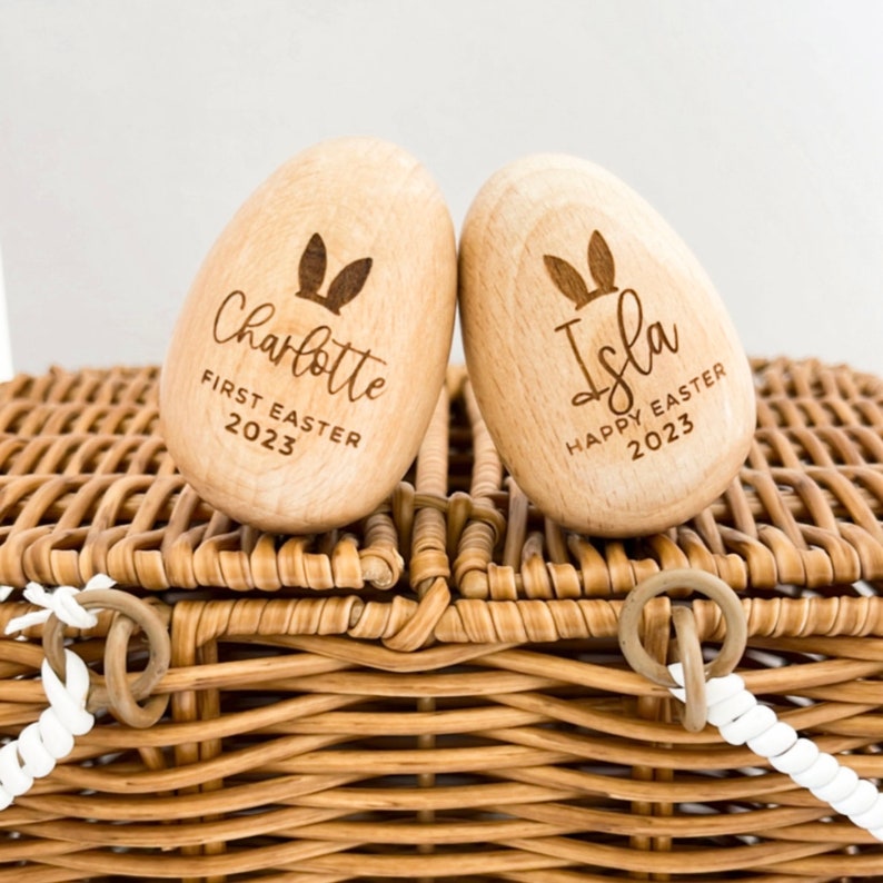May include: Two wooden Easter eggs with bunny ears engraved on them. One egg says "Charlotte First Easter 2023" and the other says "Isla Happy Easter 2023".