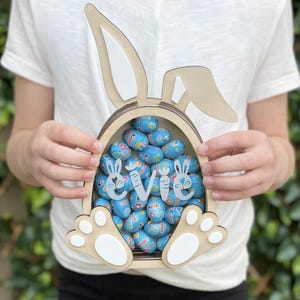 May include: A wooden Easter bunny-shaped basket filled with blue foil-wrapped chocolate eggs. The basket features bunny ears and paw-shaped feet. The name "Evie" is written on the front in white script. The basket is held by a person wearing a white t-shirt.