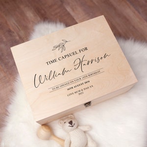 May include: A wooden time capsule box with the text "TIME CAPSUEL FOR William Harrison" and other text. The box is closed and sits on a white fluffy surface. A small teddy bear and rattle are in the foreground.