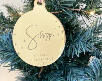 May include: Gold Christmas ornament with the name Sienna and the text "My First Christmas 2023".
