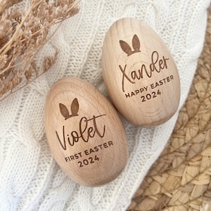 May include: Two wooden Easter eggs with bunny ears engraved on them. One egg says "Violet First Easter 2024" and the other says "Xander Happy Easter 2024".