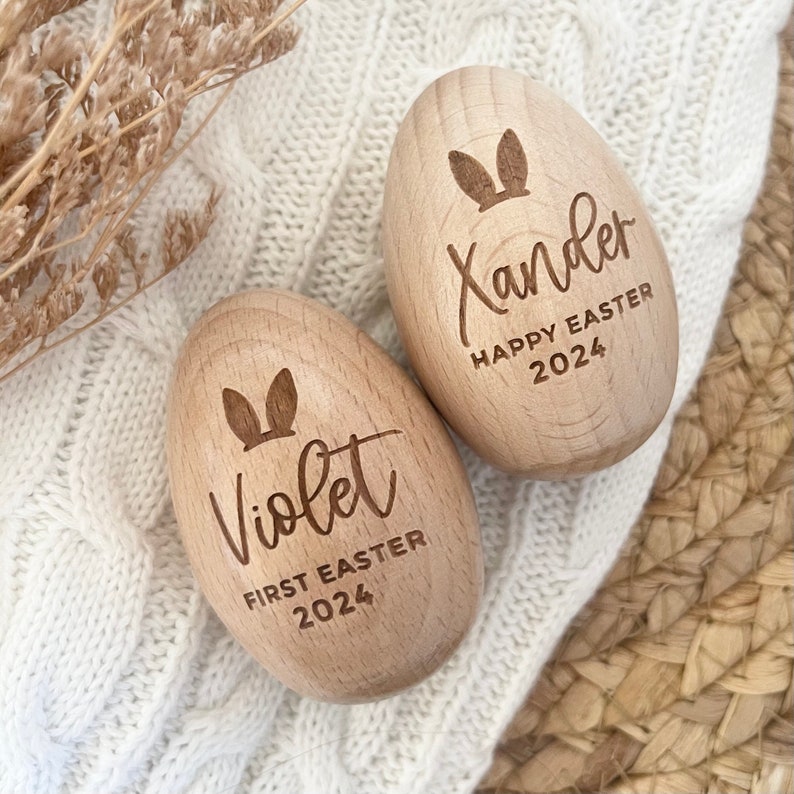 May include: Two wooden Easter eggs with bunny ears engraved on them. One egg says "Violet First Easter 2024" and the other says "Xander Happy Easter 2024".