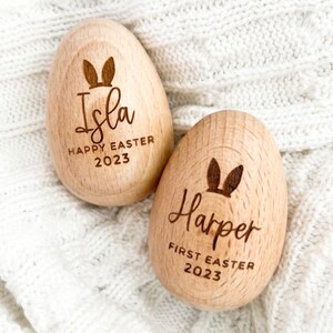 May include: Two wooden Easter eggs with bunny ears engraved on them. One egg says "Isla Happy Easter 2023" and the other says "Harper First Easter 2023".