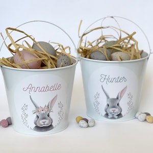 May include: Two white metal buckets with bunny rabbit illustrations and personalised names, Annabelle and Hunter. Each bucket is filled with straw and pastel coloured Easter eggs.
