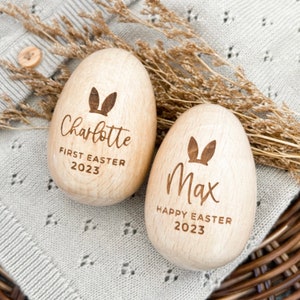 May include: Two wooden Easter eggs with bunny ears engraved on them. One egg says "Charlotte First Easter 2023" and the other says "Max Happy Easter 2023".