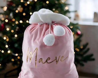 May include: A light pink velvet gift sack featuring a white faux fur cuff at the top, two white pom-poms on a drawstring cord, and the name Maeve printed in a metallic gold script font across the center of the fabric surface.