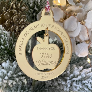 May include: Gold mirrored Christmas ornament with the text "IT TAKES A BIG HEART TO HELP SHAPE LITTLE MINDS" around the edge. Inside the circle is a cutout of an apple with the text "THANK YOU Mrs Williams" and "Love Amelia 2023" below.