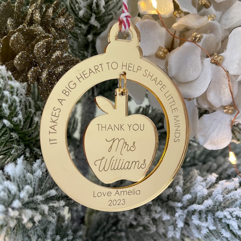 May include: Gold mirrored Christmas ornament with the text "IT TAKES A BIG HEART TO HELP SHAPE LITTLE MINDS" around the edge. Inside the circle is a cutout of an apple with the text "THANK YOU Mrs Williams" and "Love Amelia 2023" below.